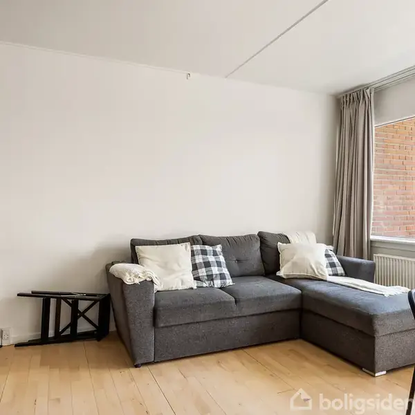 En grå hjørnesofa med puder står i en lys stue. Ved siden af er et træbord med en stol. Store vinduer viser bygninger udenfor. En hvid væg og et lille, sort bord ses også.