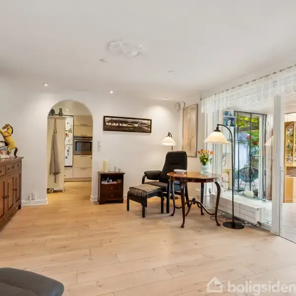 Stue med trægulv, sofa og bord ved glasdør til udestue. Reol med dekorationer, vægmonteret TV og lyskilder skaber en hyggelig atmosfære. Blomster på bordet ved siden af en stol.