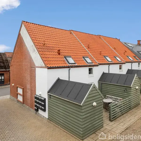 Rød murstensbygning med rødt tegltag står langs en vej. Små haveafskærmninger i trægrøn stikker ud fra siden. Skyfri blå himmel over ejendommen. Kontakteadresser er skrevet på et skilt.