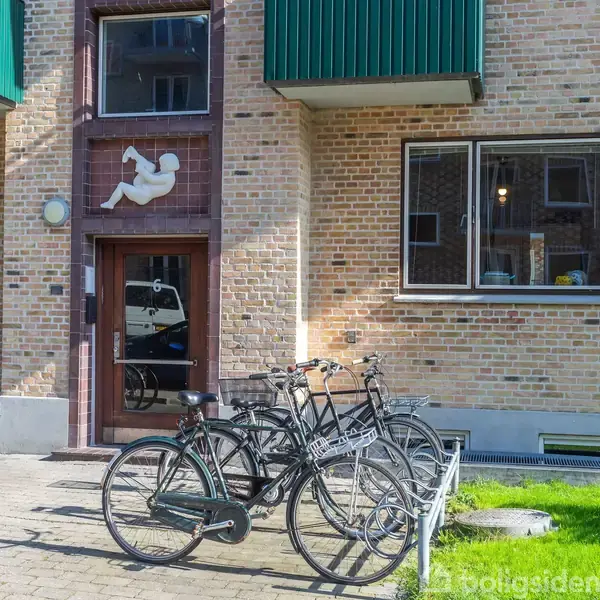 Cykler står parkeret foran en murstensbygning med en dekoration af en baby på facaden. Bygningen har store vinduer og en græsplæne ved siden af fortovet.