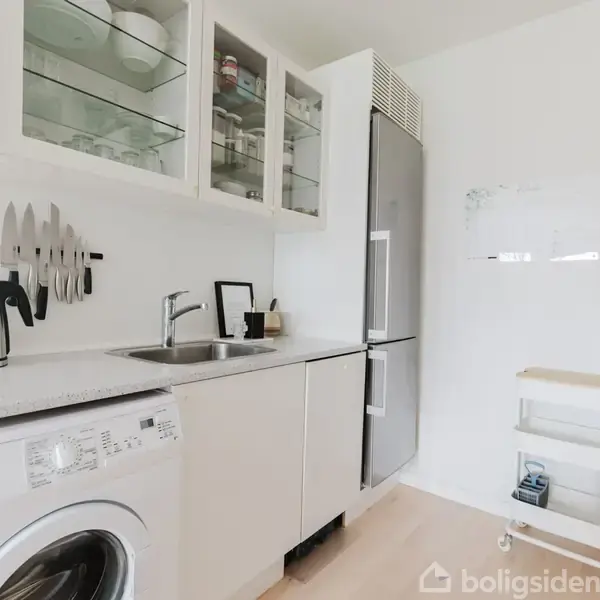 Vaskemaskine står i et lyst køkken ved siden af en kogeplade og elkedel. Knive hænger på væggen over vasken. Køleskab til højre, hylder med glas låger over vasken.