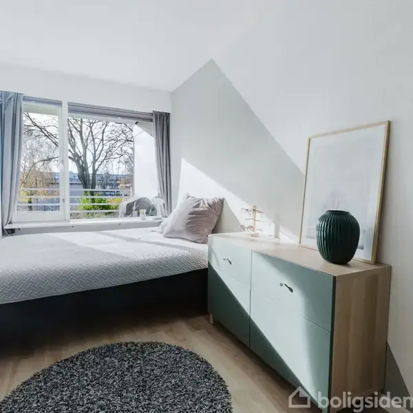 Seng med puder står ved store vinduer i et lyst værelse. Ved siden af står et skab med en vase og en ramme. Udsigt til træer udenfor. Peberkorn-formet tæppe på gulvet.