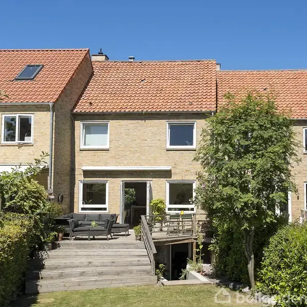 Rækkehus i mursten har rødt tegltag. Stor træterrasse med havemøbler foran huset. Omgivet af frodig have med buske og træer.