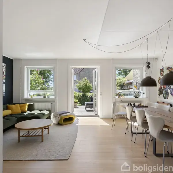Stue med grøn sofa og rundt sofabord. På bordet ved siden af sofaen ses planter. Spisestuebord med stole står under en lampe. Dør åbner ud til terrasse og have.