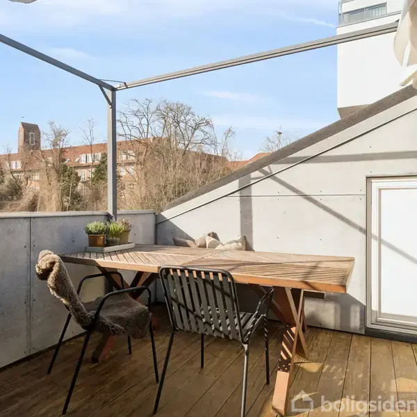 Terrassebord med stole står under en pergola. Planten på bordet giver et hyggeligt præg. I baggrunden ses et kirketårn og træer. Sollys skinner på trædækket.