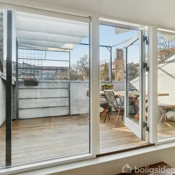 Terrasse med glasdøre åbner ud til en trædækket balkon. Et bord og stole er placeret udenfor, omgivet af bylandskab med en bygning i baggrunden under en klar blå himmel.