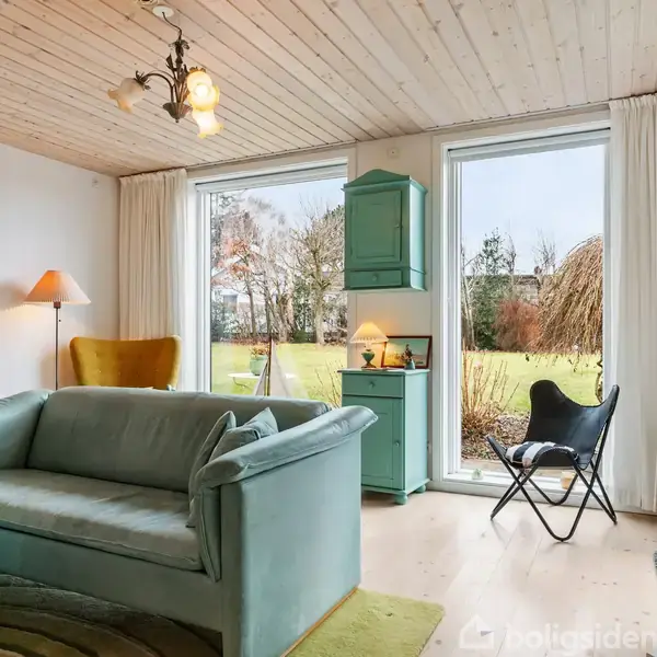 En grøn sofa står i en stue med et trægulv og lyst loft, ved siden af et skab og store vinduer, der viser en have. En dansk flag står til højre.