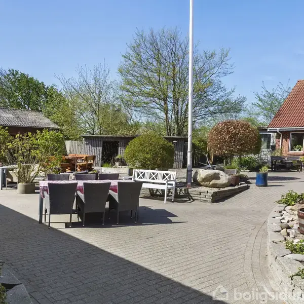 En gårdhave med havemøbler står på en brolagt terrasse. Rundt omkring er der buske, træer og røde teglsten bygninger, der tilføjer en hyggelig, landlig atmosfære.