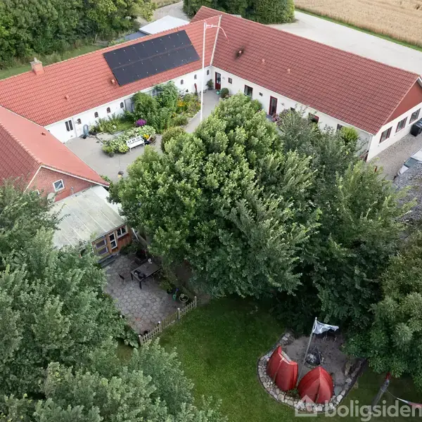 En gård set oppefra med røde tegltag og solpaneler, omgivet af træer. I gårdspladsen ses terrasse og grønt område, mens en tipi-lignende struktur står i baghaven nær en flagstang.