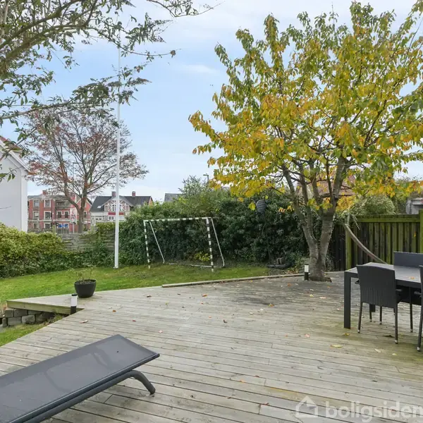 Terrasse med liggestol og spisebord, omgivet af grønne træer og buske. En lille gynge ses i baggrunden ved siden af et hvidt hus.