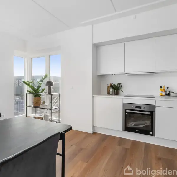 Køkken i moderne stil med hvide skabe og en indbygget ovn. Ved siden af, et spisebord og stole. Store vinduer slipper lys ind og viser en byudsigt.