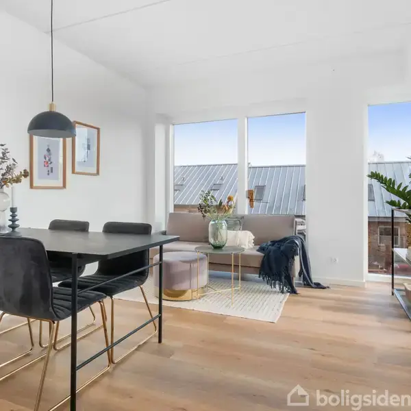 Et moderne stueområde med spisebord, fire sorte stole og hængende lampe. I baggrunden en grå sofa, to guldfarvede puffer og en plante, belyst af store vinduer.