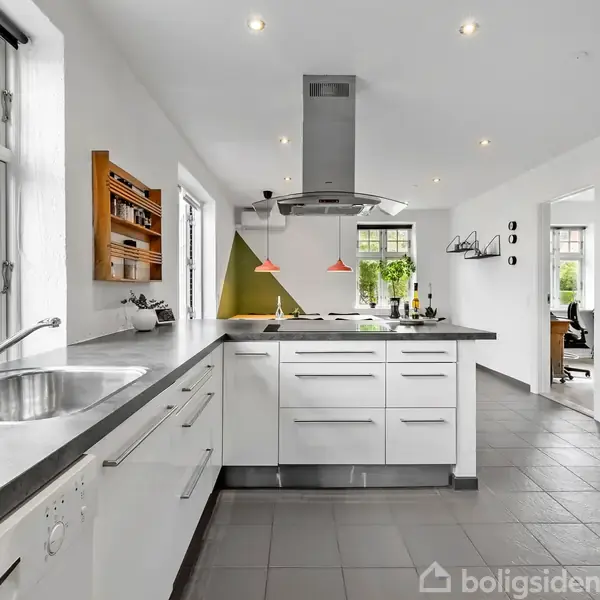 Et moderne køkken med en central ø med kogeplade og emhætte, omgivet af hvide skabe. Store vinduer tilfører lys. Til venstre ses en opvaskemaskine. Retningsskilte og dekorationer pryder væggene.