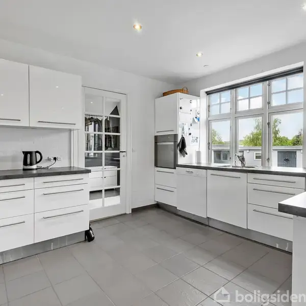 Et moderne køkken med hvide skabe og grå bordplader. En elkedel står på bordet under et stort vindue. Lysegrå fliser på gulvet. En glasdør fører til et andet rum.