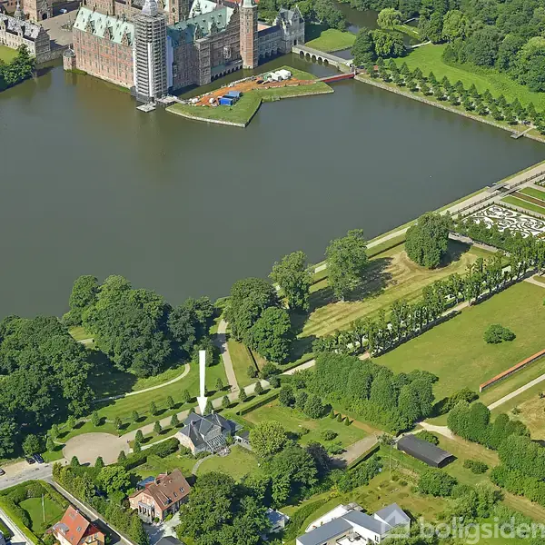 Et slot står ved en stor sø omgivet af frodig vegetation og velplejede haver. I forgrunden ses en park med græsarealer og stier, overkantet af træer og buske.