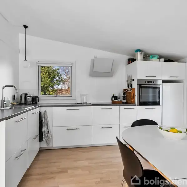 Køkken med hvide skabe og sorte håndtag. Der er en indbygningsovn, kogeplader, en vask og en skål med frugt på spisebordet. Rummet er lyst med trælaminatgulv.