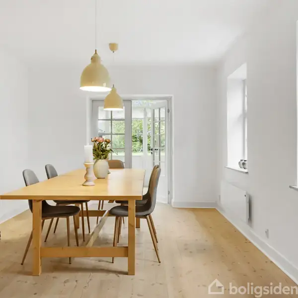 En spisebord i træ står i et lyst rum med fem stole omkring det. Der hænger to lamper over bordet. Vinduer og døre fører ud til en have.