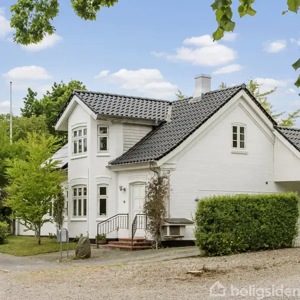Et hvidt murstenshus med sort tegltag står i en have med grønne træer. Trappen foran hoveddøren fører til indgangen. Der er vinduer med sprosser og en grussti ved siden af.