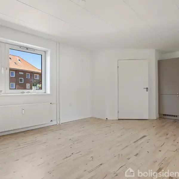 Et lyst rum med trægulv, et stort vindue viser udsigt til lejlighedsbygninger. Til højre ses et lille køkken med skabe, og en lukket dør leder til et andet rum.