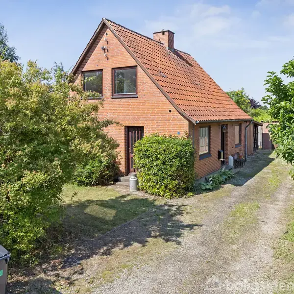 Hus i mursten med rødt tegltag, omgivet af grønne buske og træer. Stien foran huset fører til hovedindgangen. Skraldespande står til venstre ved indgangsstien.