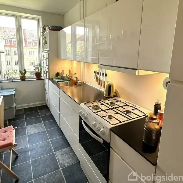 Et smalt køkken med hvide skabe og sort bordplade. En gas-komfur er aktivt med to kogende gryder. Et lille bord med stol er placeret ved vinduet, der giver udsigt til bygninger. Flasker og planter pryder hylderne.