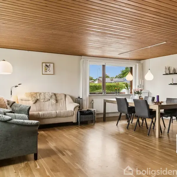 En lys stue med træloft og trægulv. To sofaer står ved et vindue, mens et spisebord med seks stole er placeret i hjørnet. Hylder pynter væggen.