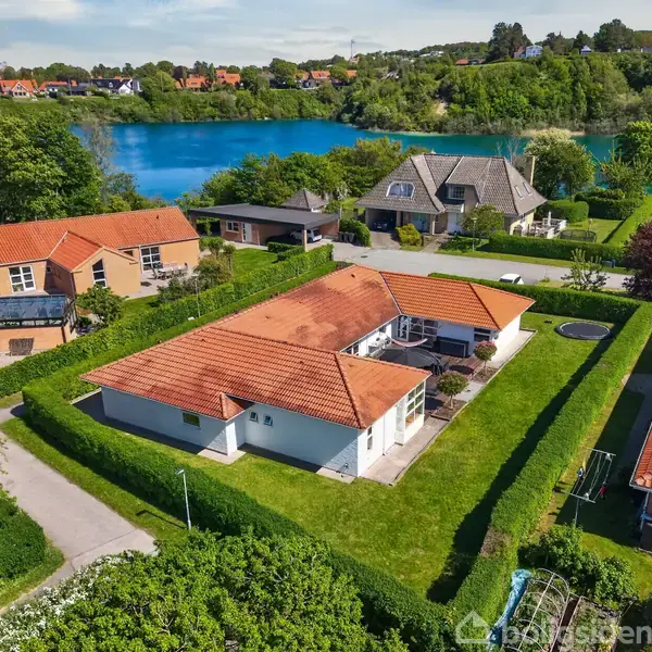 Huset med rødt tegltag ligger i en kæde af villaer, omkranset af grønne hække. Baggrunden består af en sø og en skovklædt bakke. Boligsiden-logoet er synligt.