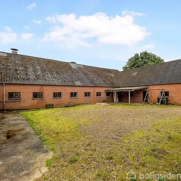 En stor, rød murstenslade står i en gårdsplads med grus og sporadisk græs. Den har et tag af bølgeeternit, og små træer omkranser stedet under en blå himmel.