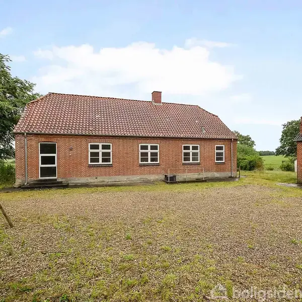 En enfamilies murstensvilla med rødt tegltag står stille i en grusbelagt gårdsplads. Omkring huset ses landlige omgivelser med grønne træer og marker i baggrunden.