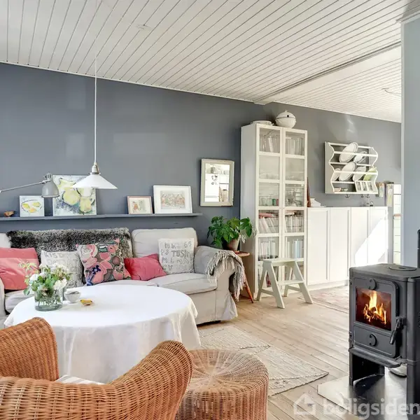 Stue med sofa omgivet af kunst, hylde med billeder, og skab i baggrunden. En rund bordplade dækket med dug har en vase med blomster. Brændeovn står til højre.