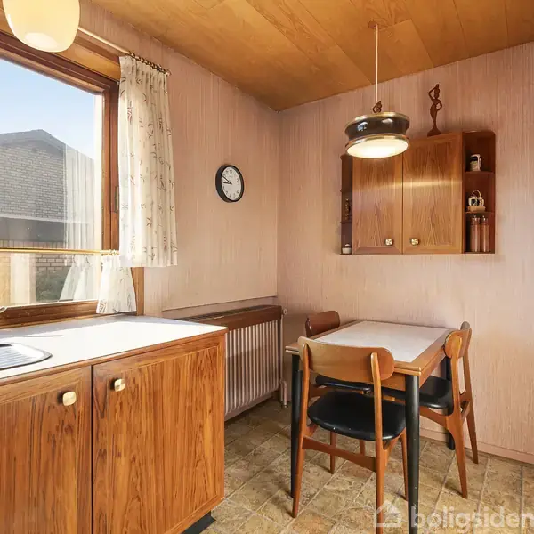 Et lille køkken med træmøbler og bord med fire stole. En lampe hænger over bordet, og et vægur hænger på væggen. Skab og hylde findes i baggrunden.
