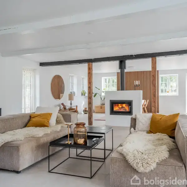 Stue med to grå sofaer og hvide tæpper, centralt placeret pejs i brug. Indrettet med træbjælker, hvide vægge, planter og store vinduer, der giver naturligt lys.