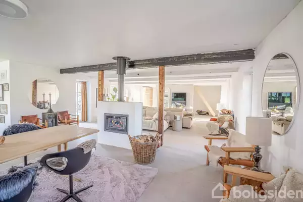 Stue med spisebord foran pejs, omgivet af moderne møbler i lyst og åbent rum. Store spejle og bjælker dekorerer væggene, mens naturligt lys skinner ind gennem vinduerne.