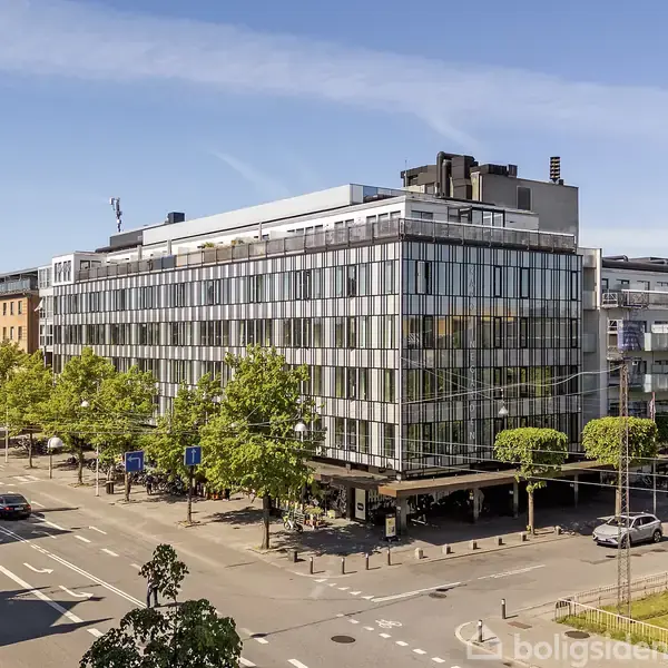 En moderne kontorbygning med glasfacade står ved en travl vej. Biler kører og træer foretager en grøn ramme omkring området under en klar blå himmel.