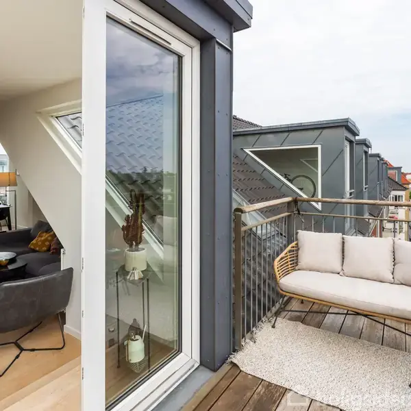 En balkon med en sofa og rundt bord i hjørnet af en moderne lejlighed med store glasvinduer, lys trægulv og udsigt over en by.