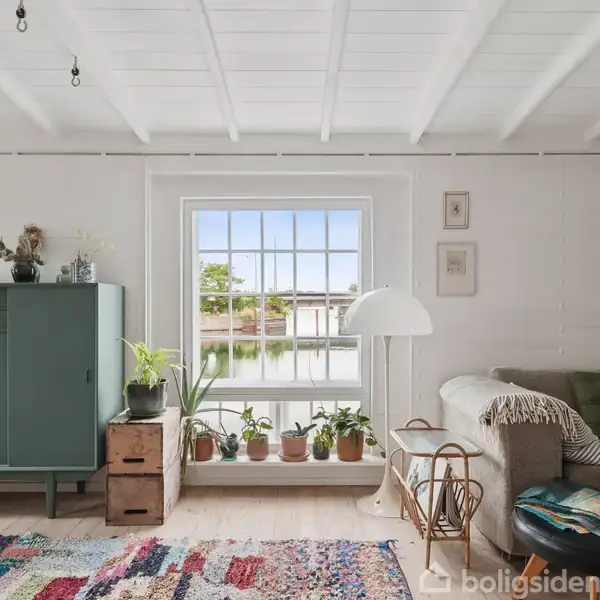 Stue med grøn kommode, planter og kvadratisk vindue, hvilket giver udsigt til udendørs natur. Farverigt tæppe ligger på gulvet. En sofa med puder står til højre.