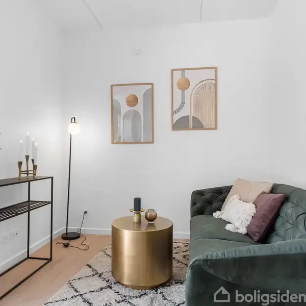 Grøn sofa med puder står ved et rundt, gyldent bord i en lys stue. En lampe og hylde med blomster står ved siden af. To abstrakte malerier hænger på væggen.