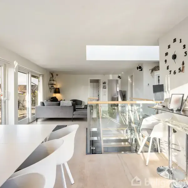 Moderne, lyst køkken-alrum med spisebord, stole og barstole ved køkkenø. Store vinduer mod venstre lader naturligt lys ind. Trappe fører nedad, og en væg har en kreativ urskive.