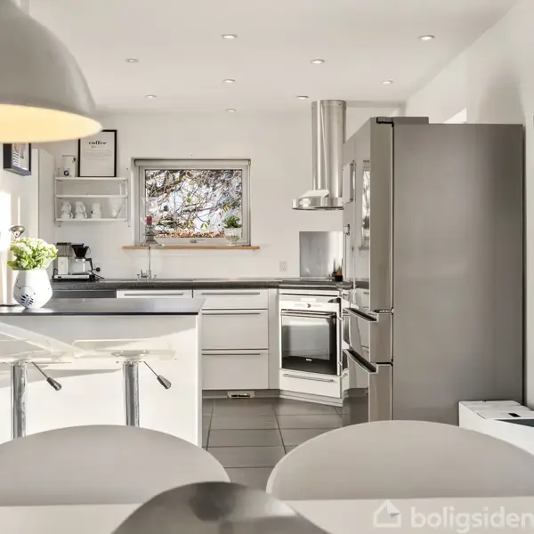 Et moderne køkken med hvide skabe og stålapparater. En stor køleskab står til højre. I forgrunden er et spisearrangement med hvide stole. Et vindue giver naturligt lys.