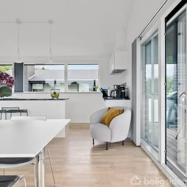 Moderne køkken-alrum med hvidt spisebord og sorte stole, blomster i vase på bordet. Skydedøre fører til terrasse med havemøbler. Lyse trægulve og vinduer mod haven giver lys.
