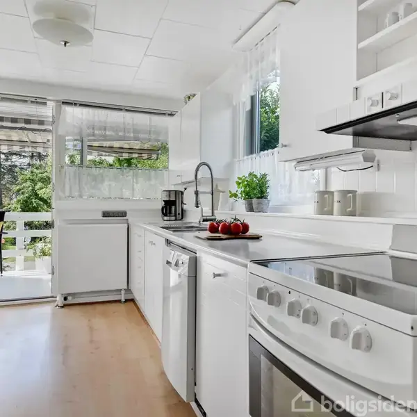 Et lyst, hvidt køkken med en komfur og vaskeområde. Bordpladen er dekoreret med rød frugt og en plante. Store vinduer giver udsigt til en frodig have.