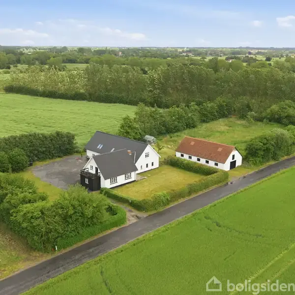 Et hvidt hus med sort tag og en mindre udbygning står stille omgivet af grønne marker og træer på en landlig vej.