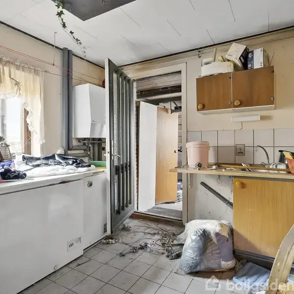 Køkkenskabene og køkkenbordet er fyldt med rod og diverse genstande. En stor kummefryser står til venstre. Gennem en dør ses en gang. Gulvet er flisebelagt.