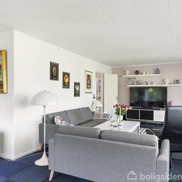 Grå sofa står i moderne stue med blå gulvtæppe. Der er et sofabord foran, omkranset af stole. Bagved står reoler med pynt, og en stor fladskærm tv. Flere vægdekorationer hænger, inklusiv et maleri med blomster.
