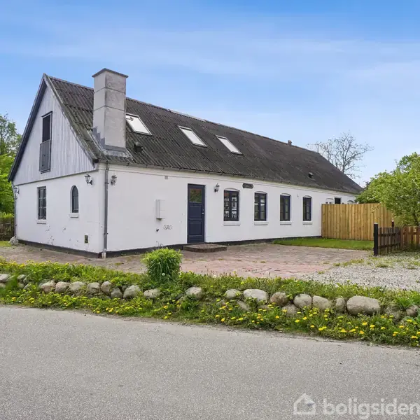 Hvidmalet hus med sort tag og flere vinduer, stående langs en vej med stenbelagt indkørsel og have omkranset af grønne træer og hegn i baggrunden.