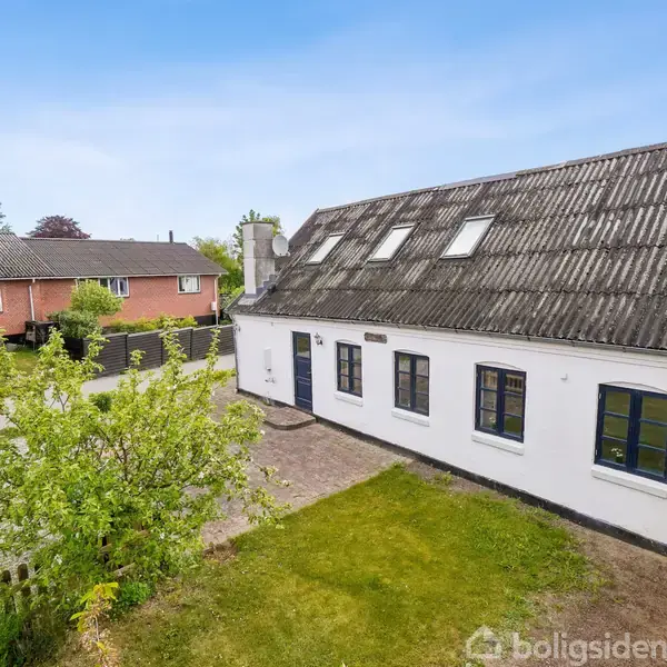 Hvidt hus med skråt sort tag står i grøn forhave. Træ og hækkens grønne blade omkranser grusvej nær rødt murstenshus. Klar blå himmel ses i baggrunden.