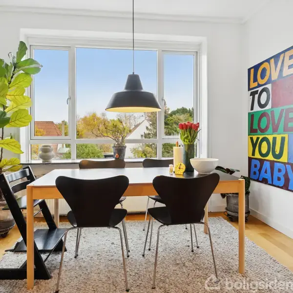 Et spisebord står i et lyst rum foran et stort vindue, med en moderne lampe hængende over. På væggen til højre hænger et farverigt skilt med teksten: "LOVE TO LOVE YOU BABY".