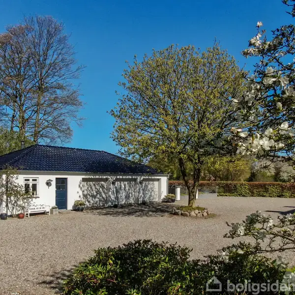 En hvid villa med mørkt tag står stille ved et grusareal omgivet af grønne træer og blomstrende buske under en klar blå himmel.