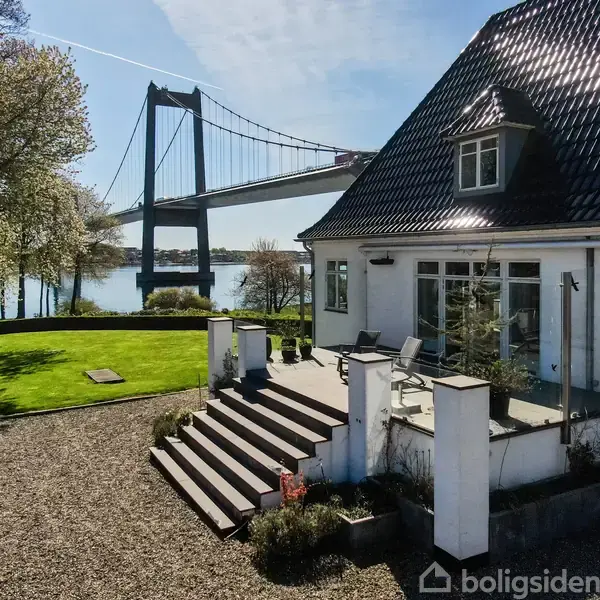 Hvidt hus med sort tag står i en have ved siden af en bro. Solen skinner på omgivelserne, og en flod løber under broen.