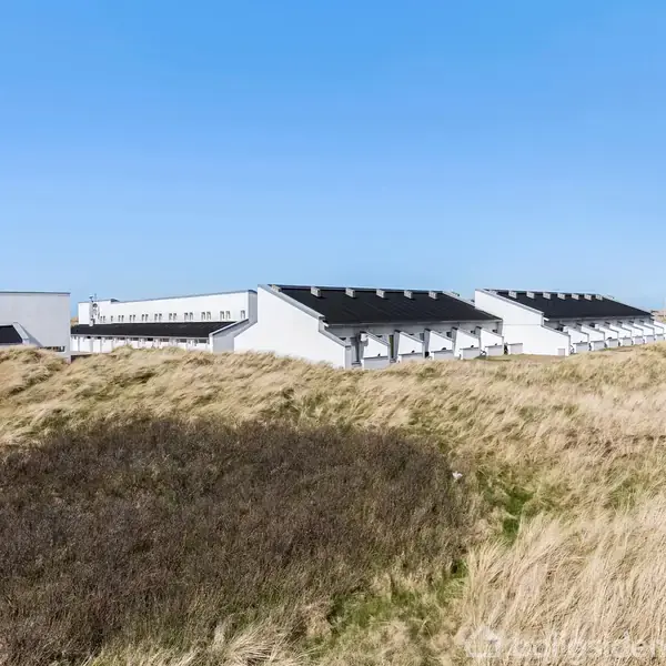 Flere ensartede, hvide rækkehuse står på en flad mark dækket af tørre græsarealer under en skyfri blå himmel, hvilket giver indtryk af en fredelig og afsidesliggende boligområde.